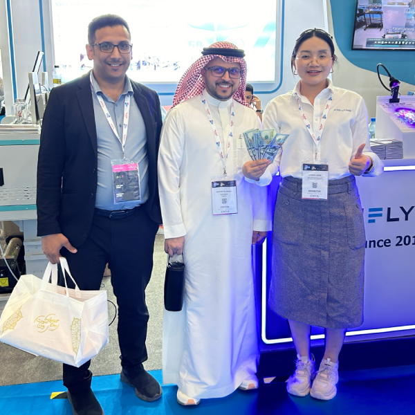 A crowded trade show booth featuring a Direct to Film printer as the centerpiece. The company team is gathered for a group photo while attendees explore printed garments and materials around them.